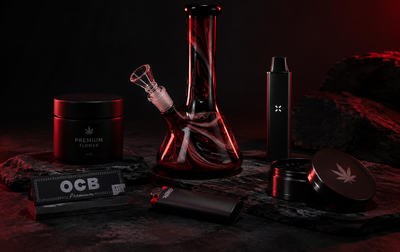 Glass water pipe, papers, grinder and vape on a dark slate surface